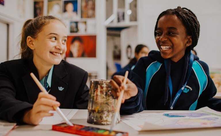 Two senior school pupils smiling and painting at St Margaret's School Bushey