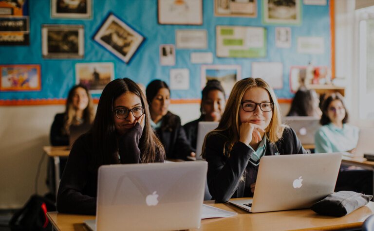 Two St Margaret's School sixth form girls smiling and talking working on laptops in classroom