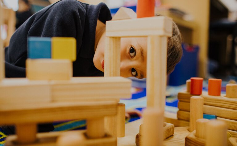 Nursery pupil peeking through wooden building blocks following the EYFS curriculum at St Margaret's School Bushey