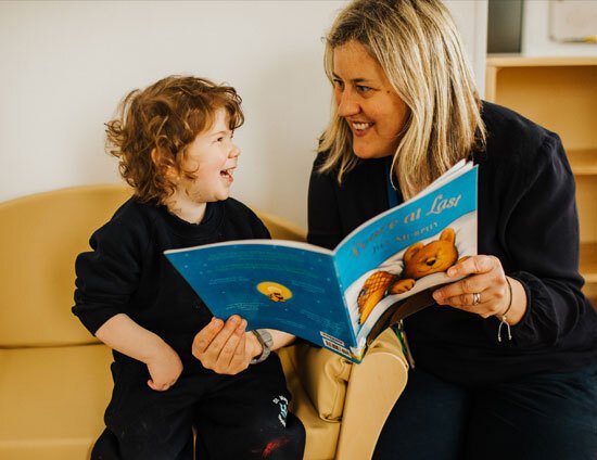 Nursery staff member reading a book with a very happy young nursery school pupil